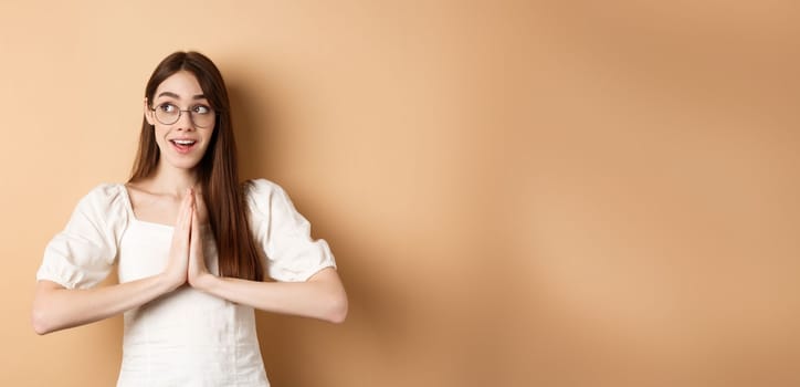 Hopeful girl holding hands in pray, begging for dream come true, looking aside excited, standing on beige background. Copy space