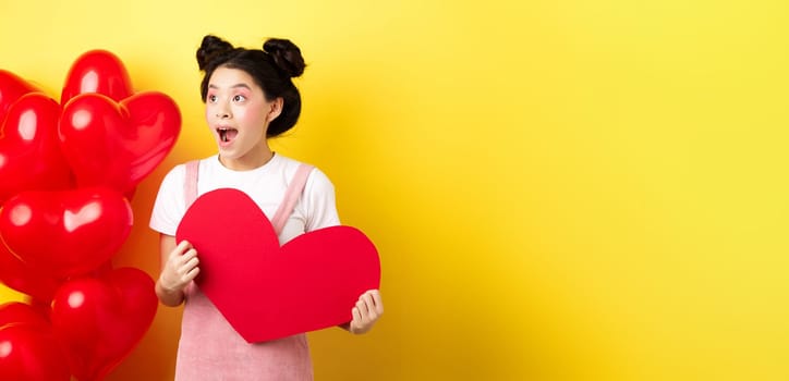 Happy Valentines day. Excited and surprised asian girl checking out romantic offer, looking left cheerful, showing big red heart card, yellow background.