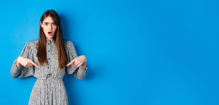 Shocked and offended young woman in dress frowning, gasping and pointing fingers down at insulting promo, standing on blue background.