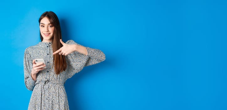 Stylish caucasian woman in dress, pointing at smartphone and smiling, showing online promo, standing on blue background.