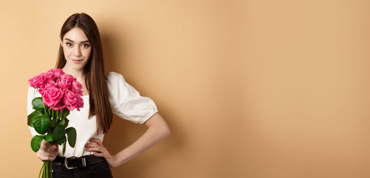 Valentines day. Beautiful girlfriend holding pink roses and looking at camera. Young woman receive flowers from her date, standing on beige background.