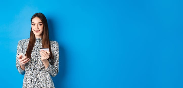 Online shopping. Happy smiling woman buying in app, holding smartphone and plastic credit card, blue background.