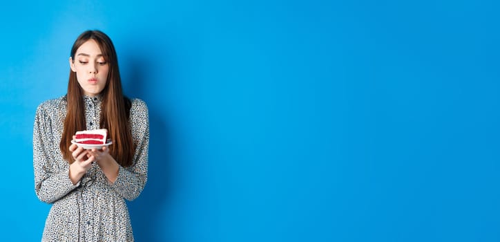 Cute birthday girl celebrating, blowing candle on cake and making wish, standing on blue background. Party and holidays concept.