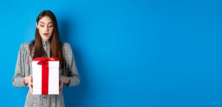 Holidays. Surprised young woman looking at gift with amazed face, receive Valentines day present, standing in dress on blue background.