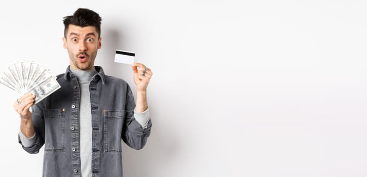 Excited guy holding plastic credit card and dollar bills, standing amused on white background.