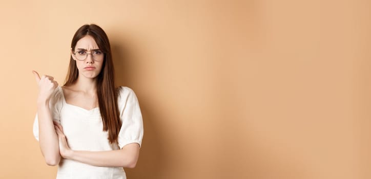 Disappointed girl in glasses frowning, pointing aside at bad product, disapprove and dislike something, standing on beige background.