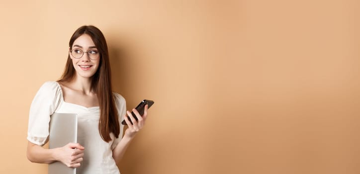 Image of young stylish woman going on work, holding laptop and smartphone, looking aside at empty space, standing on beige background.
