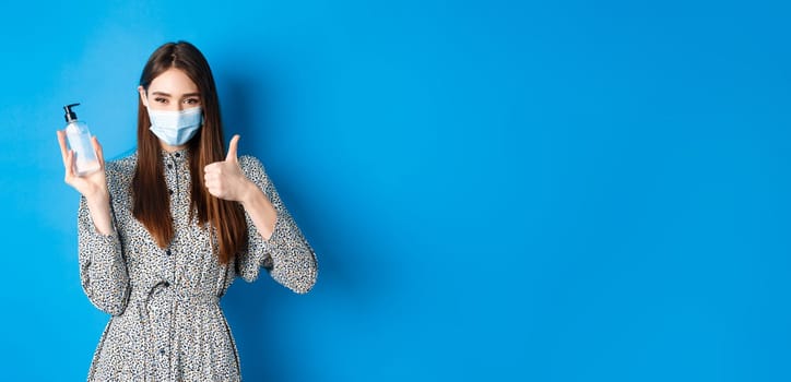 Covid-19, social distancing and healthcare concept. Beautiful girl in medical mask using hand saniziter, holding antiseptic bottle and showing thumbs up, standing on blue background.