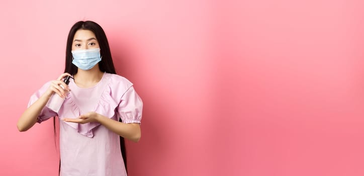 Healthy people and covid-19 pandemic concept. Cute asian woman in medical mask clean hands with antiseptic, using sanitizer, standing against pink background.