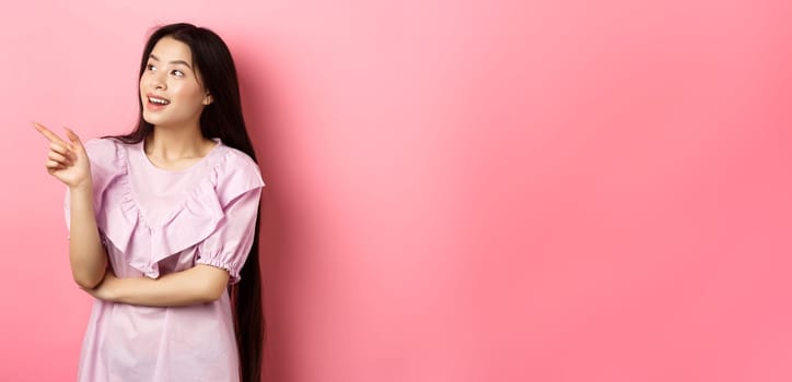 Excited smiling asian woman in dress, pointing and looking left at logo, checking out promotion, standing on pink background.