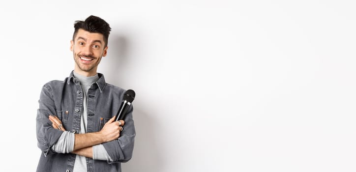 Handsome caucasian guy with moustache cross arms on chest, holding mic and smiling at camera, perform on stage with microphone, standing against white background.