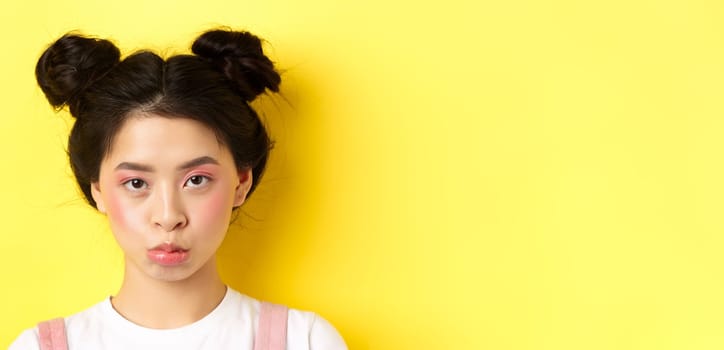 Close up portrait of moody asian girl pouting and looking upset at camera, standing wiht glamour makeup and hairbuns on yellow background.