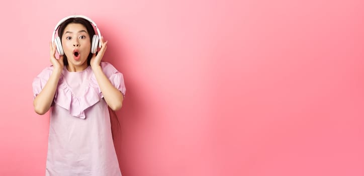 Excited asian girl hear awesome song, listen music in headphones and look amazed, standing on pink background.