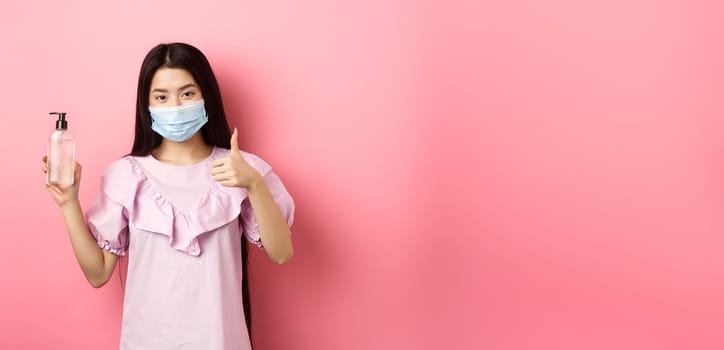 Healthy people and covid-19 pandemic concept. Cheerful asian woman in medical mask recommend hand sanitizer, showing thumb-up, praise antiseptic bottle, white background.