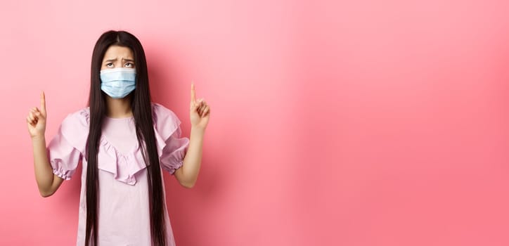 Coronavirus, quarantine and lifestyle concept. Disappointed sad asian girl pointing fingers up, frowning and sulking at unfair situation, standing against pink background.