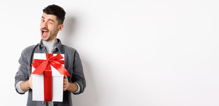 Funny young man holding valentines gift and winking at camera, making romantic surprise to lover, standing on white background.