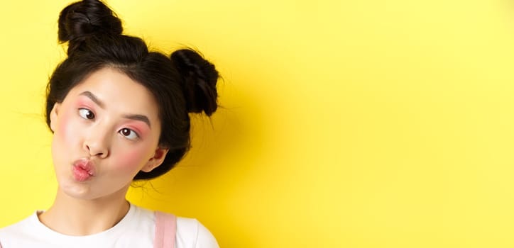 Close up portrait of funny asian teen girl with glamour makeup and hairstyle, squinting eyes and pucker lips silly, standing on yellow background.