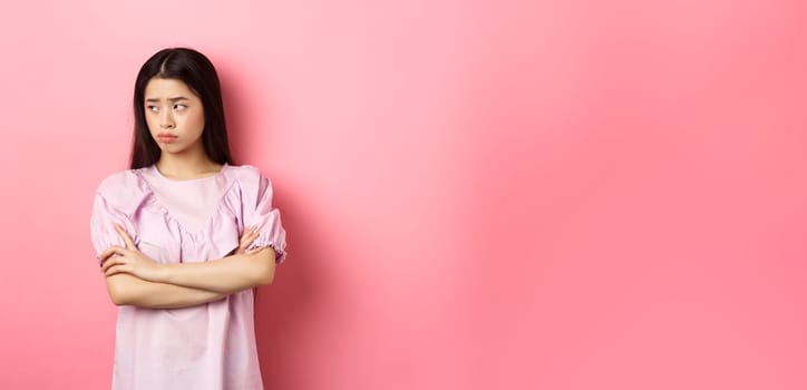 Sad and gloomy asian woman look away at logo, sulking from unfair situation, cross arms on chest disappointed, standing against pink background.