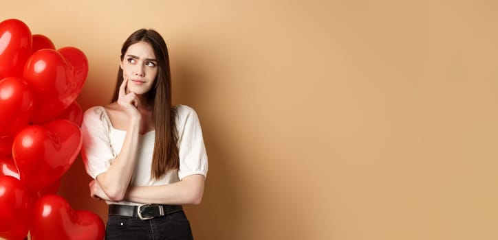 Valentines day and love concept. Pensive woman in trendy clothes and makeup, thinking and frowning, looking aside doubtful, standing near heart balloons, beige background.