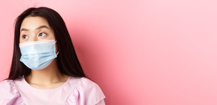 Covid-19, pandemic and quarantine concept. Close-up of dreamy asian girl in medical mask looking left at logo with thoughtful face, standing on pink background.