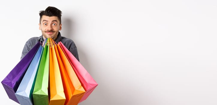 Excited young man carry colorful shopping bags and smiling happy, shopper buying on sale, standing on white background.