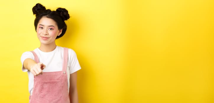 Stylish japanese girl with beauty makeup pointing at camera, smiling at you, beckon and invite people, standing on yellow background.