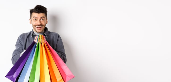 Excited smiling guy holding colorful shopping bags and rejoicing with discounts in store, standing against white background.