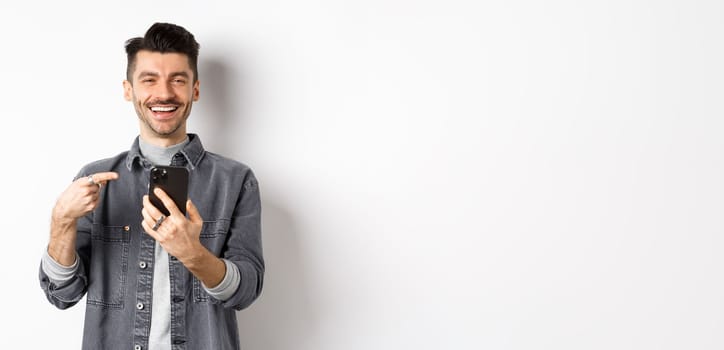 Happy handsome caucasian man with moustache pointing finger at smartphone, smiling and showing good deal online on mobile phone, standing pleased against white background.