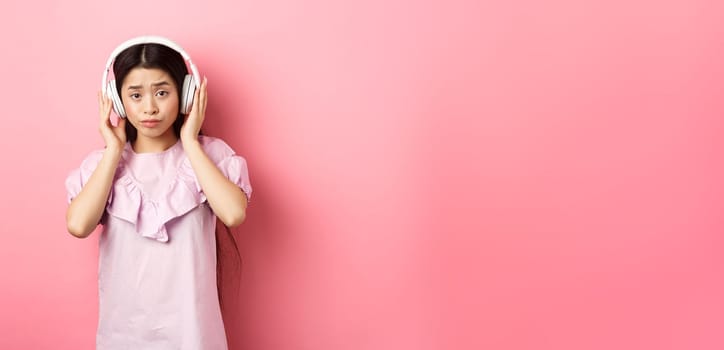 Skeptical girl unamused with song, listening music in headphones and frowning displeased, standing against pink background.