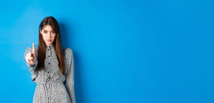 Serious woman looking confident and shaking finger, say no, stop you, disagree and disapprove, standing in dress on blue background.