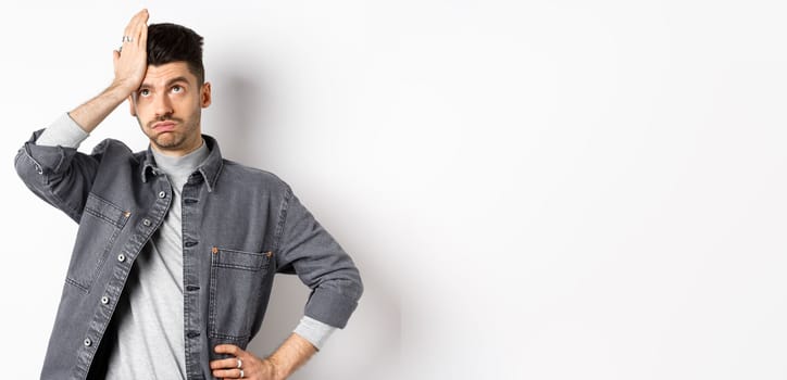 Annoyed young man looking tired or bothered, roll eyes and slap forehead, making facepalm from something stupid, standing on white background.