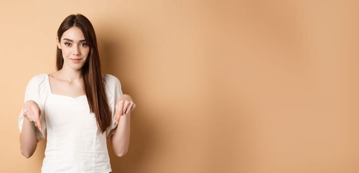 Smiling tender girl in dress pointing fingers down, showing advertisement on empty space, beige background.