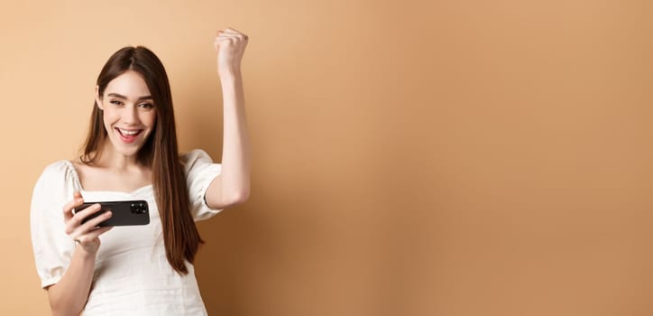 Girl winning on mobile phone. Happy woman raising hand up and scream yes with joy, achieve goal in smartphone app, standing on beige background.