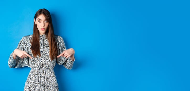 Excited beautiful woman in dress say wow, pointing fingers down and look amused, checking out promotion, standing on blue background.