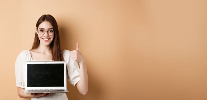 Smiling young woman in glasses showing laptop screen deal and thumb up, recommending offer, standing on beige background.