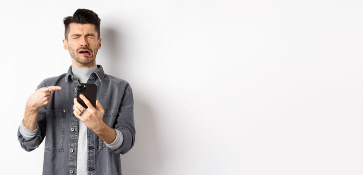 Sad crying guy pointing at smartphone and sobbing, complaining or feeling miserable, standing on white background.