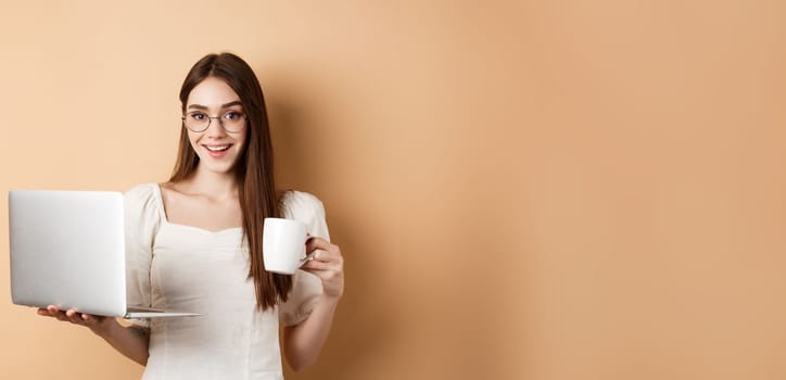 Cheerful woman in glasses drinking coffee and working on laptop, smiling at camera, standing on beige background.