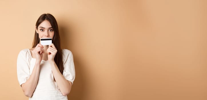 Excited girl holding plastic credit card on lips and smiling, thinking of shopping, going to buy something, standing on beige background.