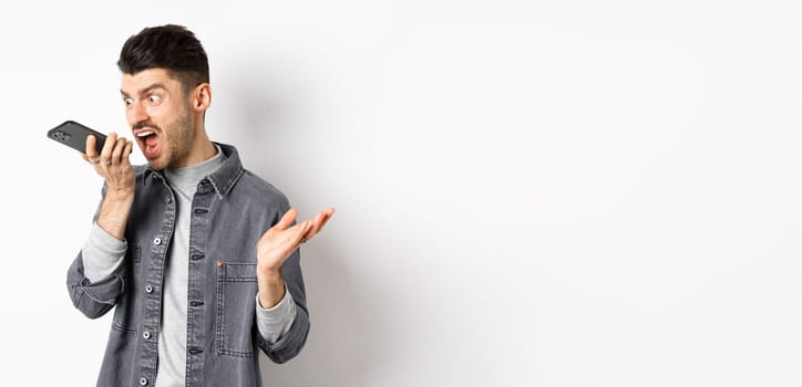 Angry guy shouting at speakerphone, scream on mobile phone, leaving angry voice message, standing against white background.