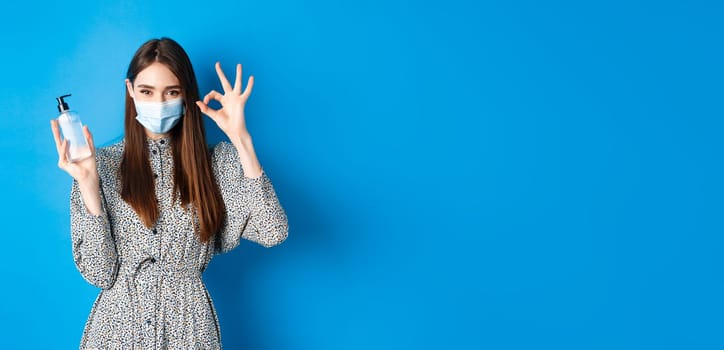 Covid-19, social distancing and healthcare concept. Beautiful girl in medical mask showing bottle antiseptic or hand sanitizer, make okay sign, recommend product, standing on blue background.