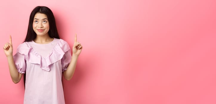 Beautiful smiling asian teen girl pointing fingers up, looking at advertisement with happy face, standing on pink background.