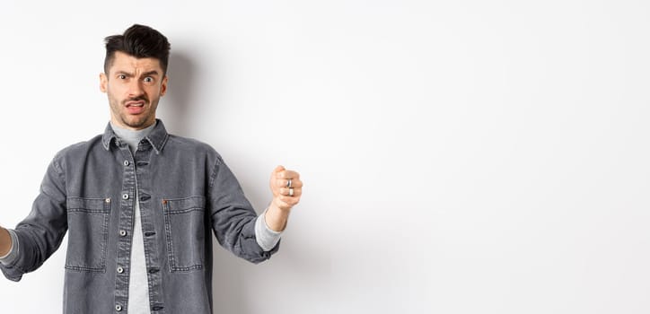 Skeptical young man showing big size object and frowning doubtful, standing reluctant on white background.