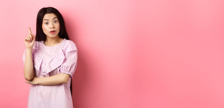 Excited asian woman pitching an idea, raising finger in eureka sign, have plan or solution, standing on pink background.