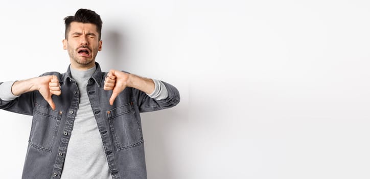 Sad man crying and showing thumbs down, whining and complainin, being upset and disappointed, standing on white background.