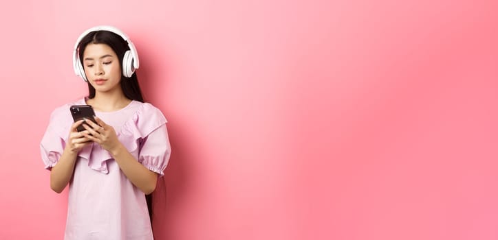 Young girl listening music in headphones and chatting on mobile phone, standing in dress against pink background.