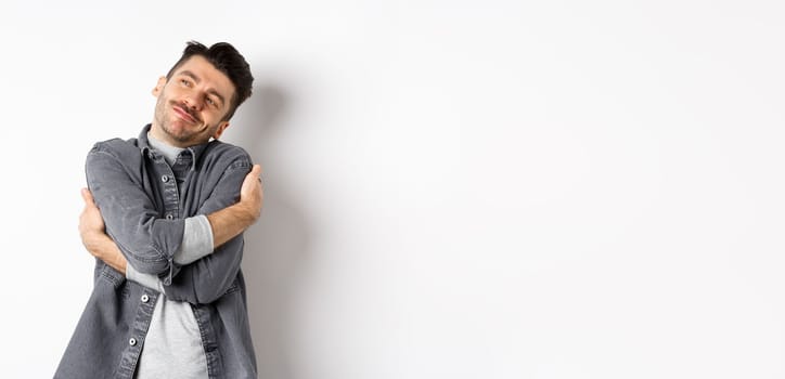 Happy romantic guy hugging himself with closed eyes and smile, loving own body, daydreaming about lover, standing on white background.