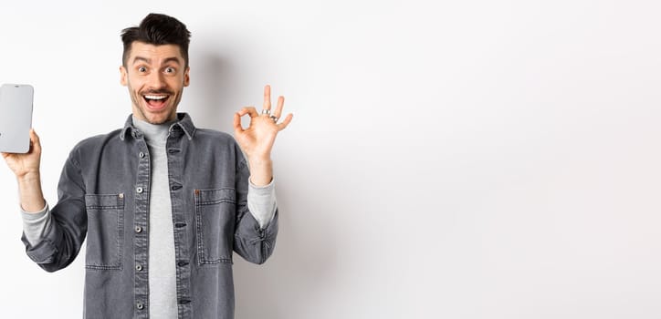 Excited smiling guy showing empty smartphone screen with okay gesture, recommending app or shopping offer, standing on white background.