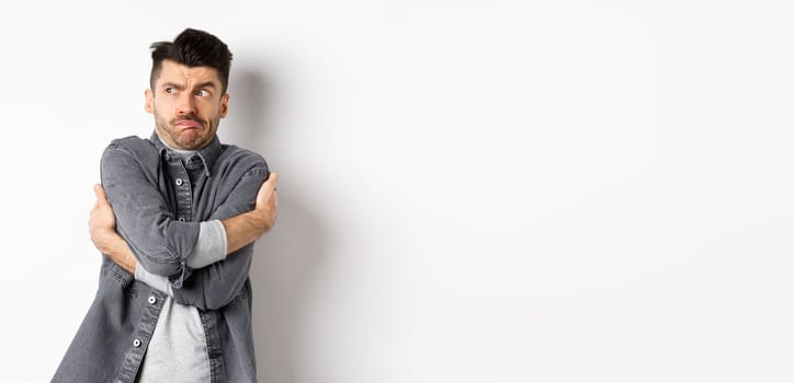 Displeased young man feeling discomfort, hugging himself and staring left at something embarrassing or exposing, standing uncomfortable on white background.