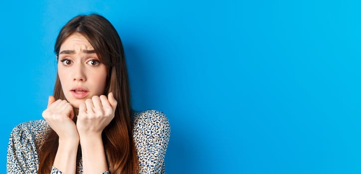 Close-up of scared timid girl looking afraid, shaking from fear, press hands to body and staring frightened, standing on blue background.