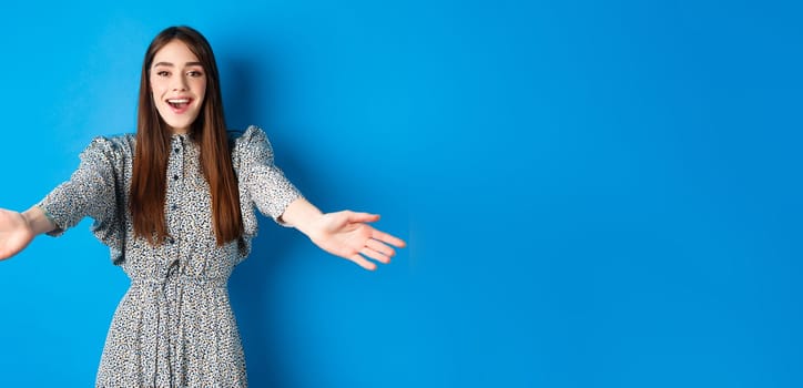 Beautiful happy woman in dress stretch out hands in warm welcome, smiling and greeting you, inviting guests, standing on blue background.